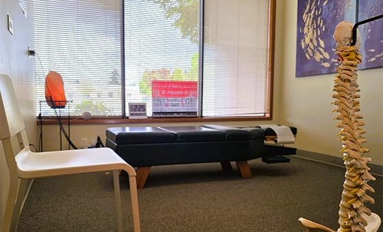 Interior of a chiropractic or therapy room featuring an adjustment table, a white chair, a model of a human spine in the foreground