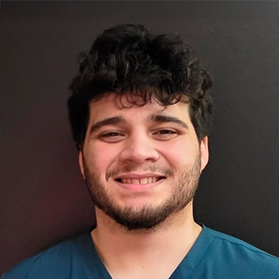 Person wearing a dark teal scrub-style shirt standing against a plain dark wall