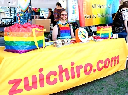 Colorful booth at an event featuring a bright yellow tablecloth with the text ‘zuichiro.com’ in pink letters, rainbow-themed decorations, and promotional items displayed on the table.