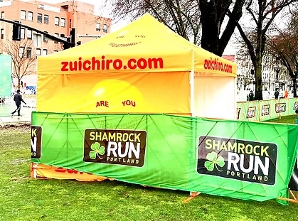 Bright yellow event tent with the text ‘zuichiro.com’ printed in red on the canopy, surrounded by green fencing featuring the Shamrock Run Portland logo with a clover design.