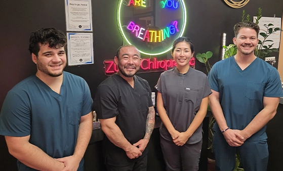 Four individuals wearing dark-colored medical scrubs standing indoors in front of a neon sign that reads ‘Are You Breathing?’ with additional text ‘Zui Chiropractic’ partially visible. The background includes framed certificates and plants