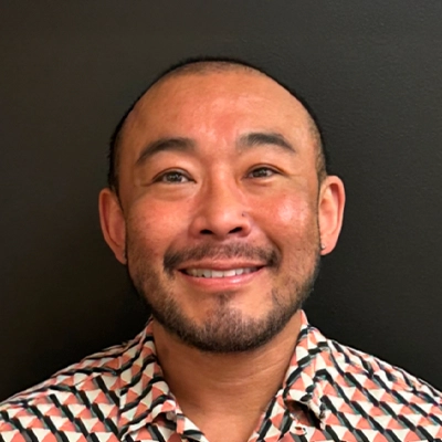 Dr Keiichi Aoyama wearing a patterned shirt with geometric shapes, standing against a plain dark background