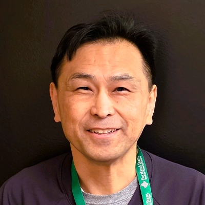 Dr Yoshifumi Ito wearing a dark top and a green lanyard with white text, standing against a plain dark wall