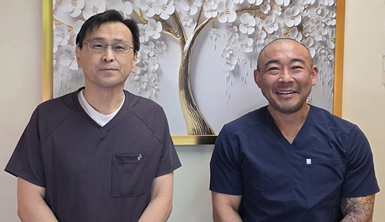 Dr. Yoshifumi Ito and Dr. Keiichi Aoyama wearing dark scrub tops seated side by side in front of a decorative wall art featuring white flowers and tree branches in a gold frame